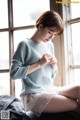 A woman sitting on a bed wearing a blue sweater and white skirt.